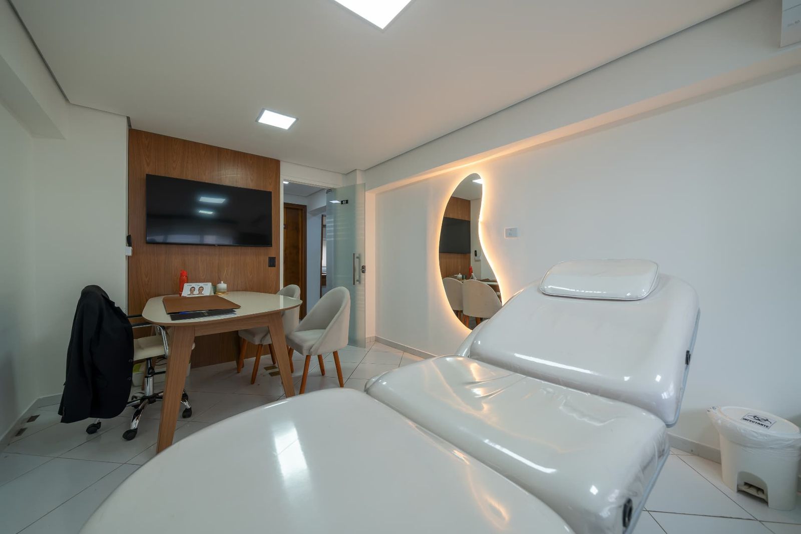 Sala com maca de estética, TV e espelho orgânico iluminado